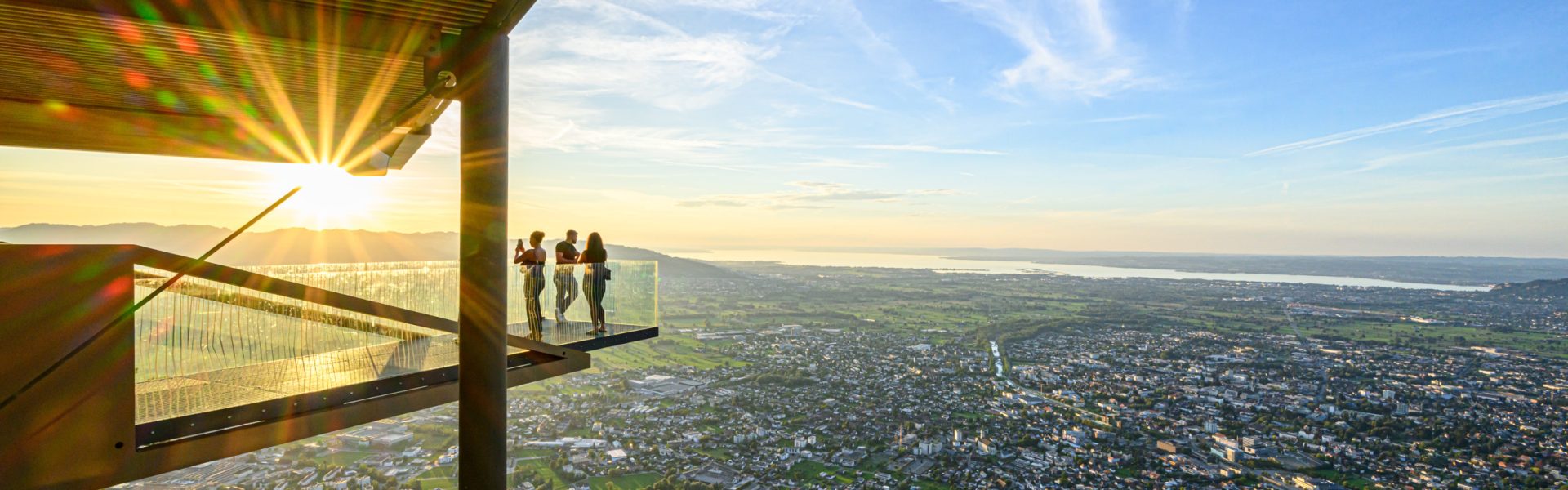 Drei Menschen genießen den Sonnenuntergang auf der gläsernen Aussichtsplattform Karrenkante auf dem Karren in Dornbirn.