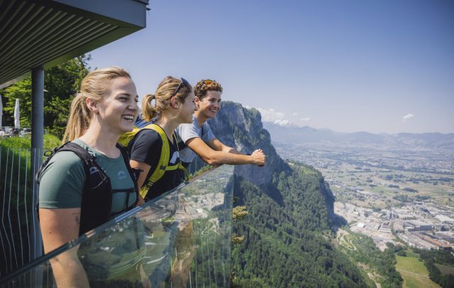 Drei Menschen genießen lächelnd die Aussicht über Dornbirn von der gläsernen Aussichtsplattform der Karrenkante auf dem Karren.